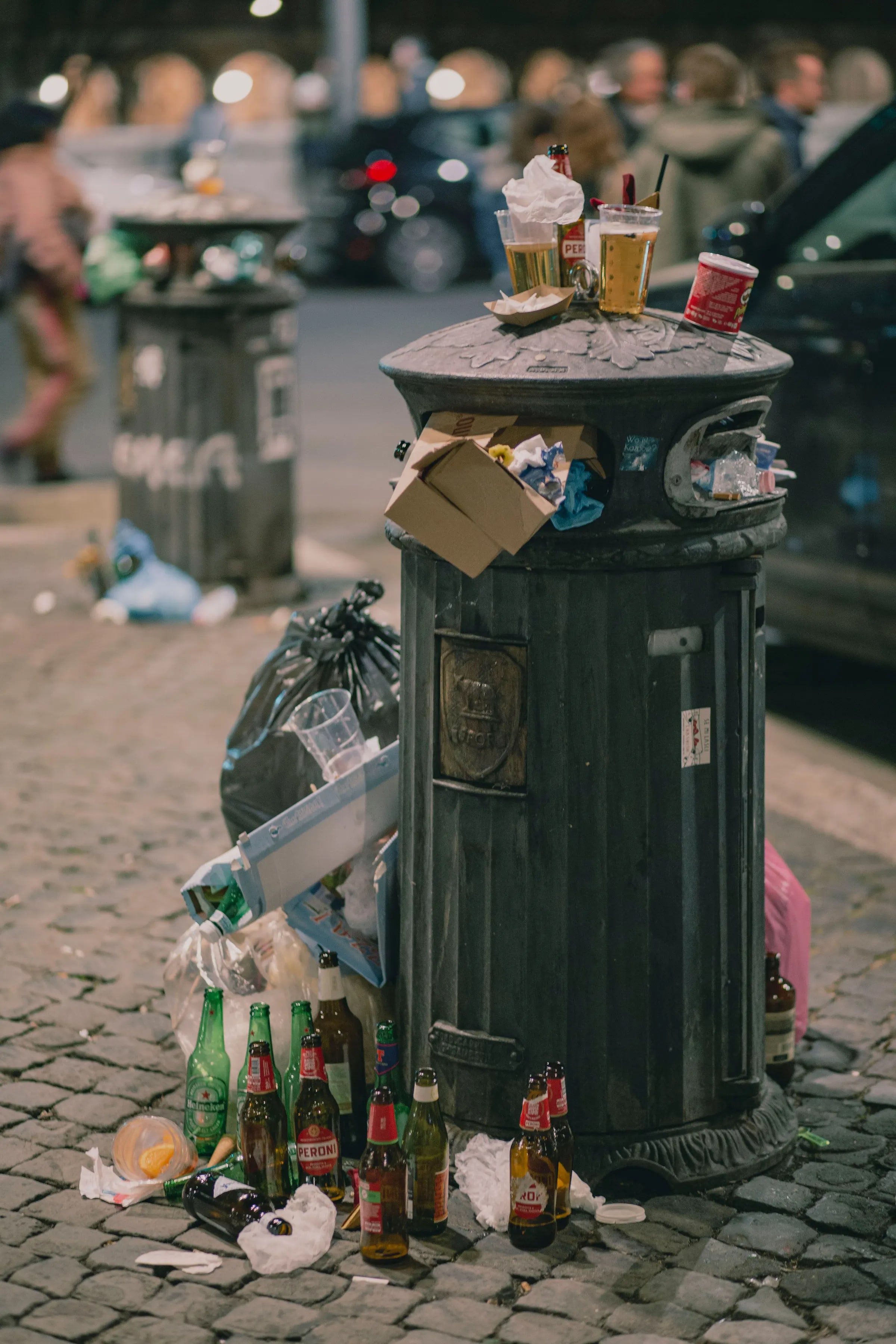 An overflowing litter bin with bottles and rubbish bags on the street