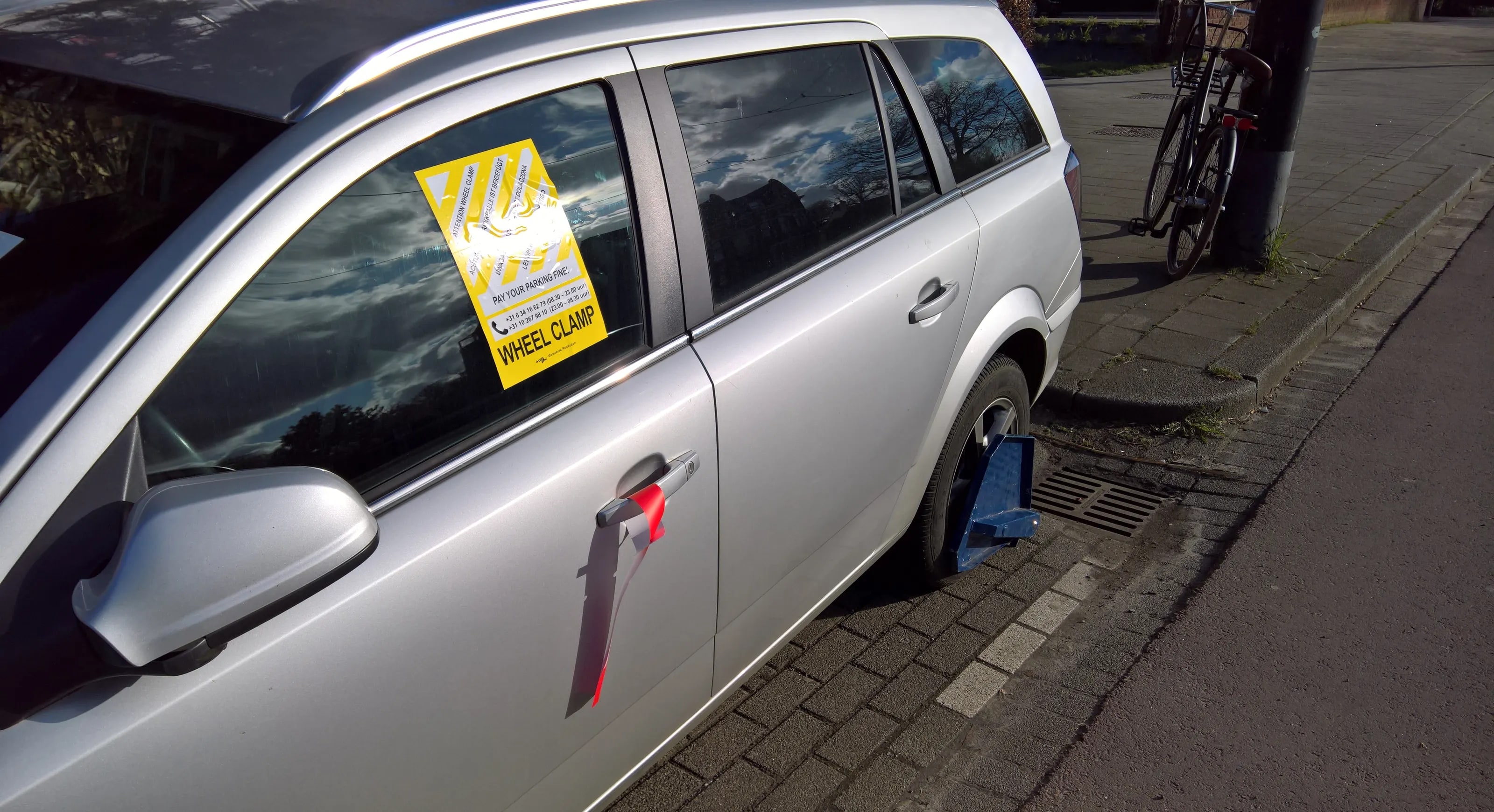 A blue wheel clamp on the rear wheel of a parked car, with the yellow notice behind the window