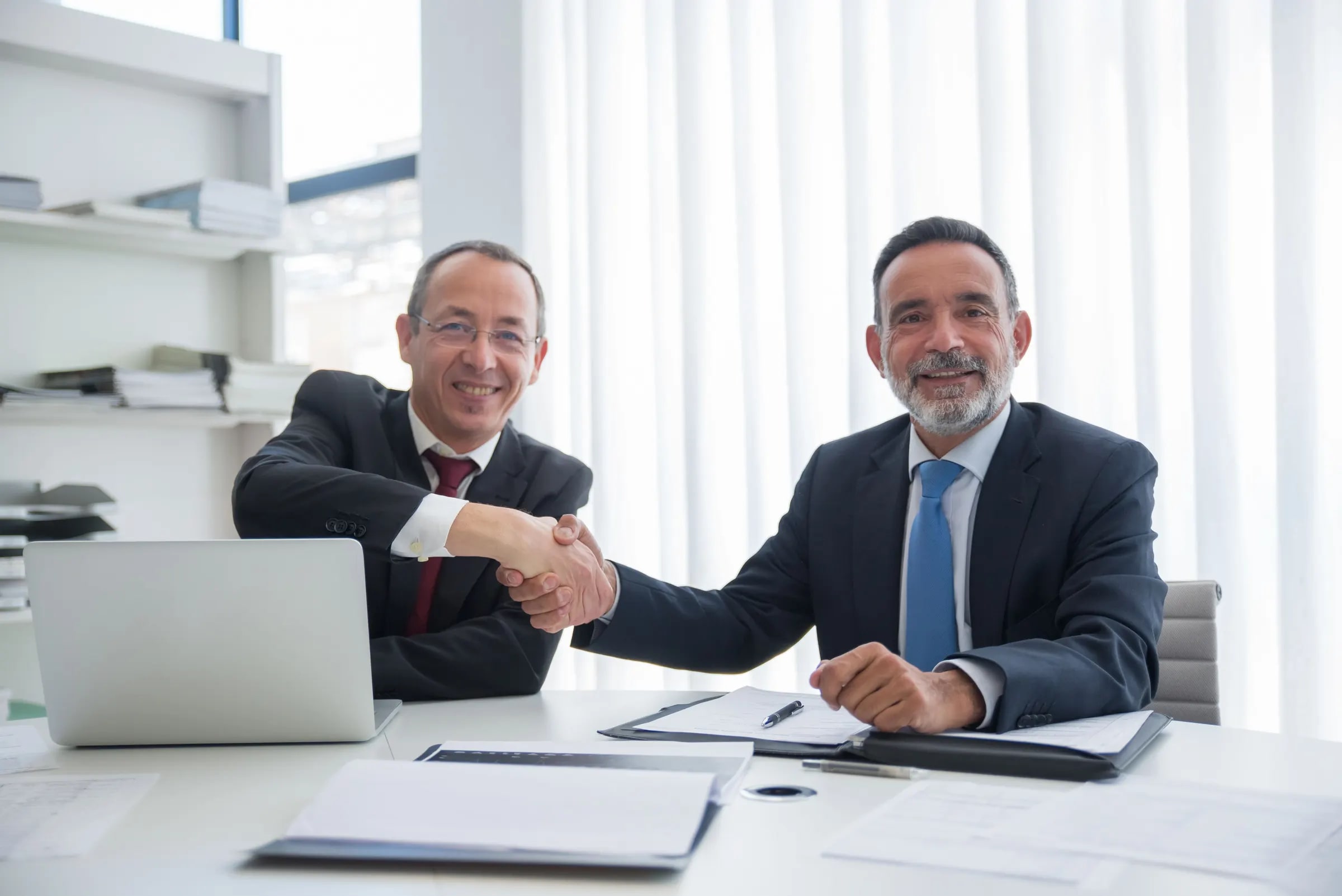Two men in suits shake hands over a desk with documents