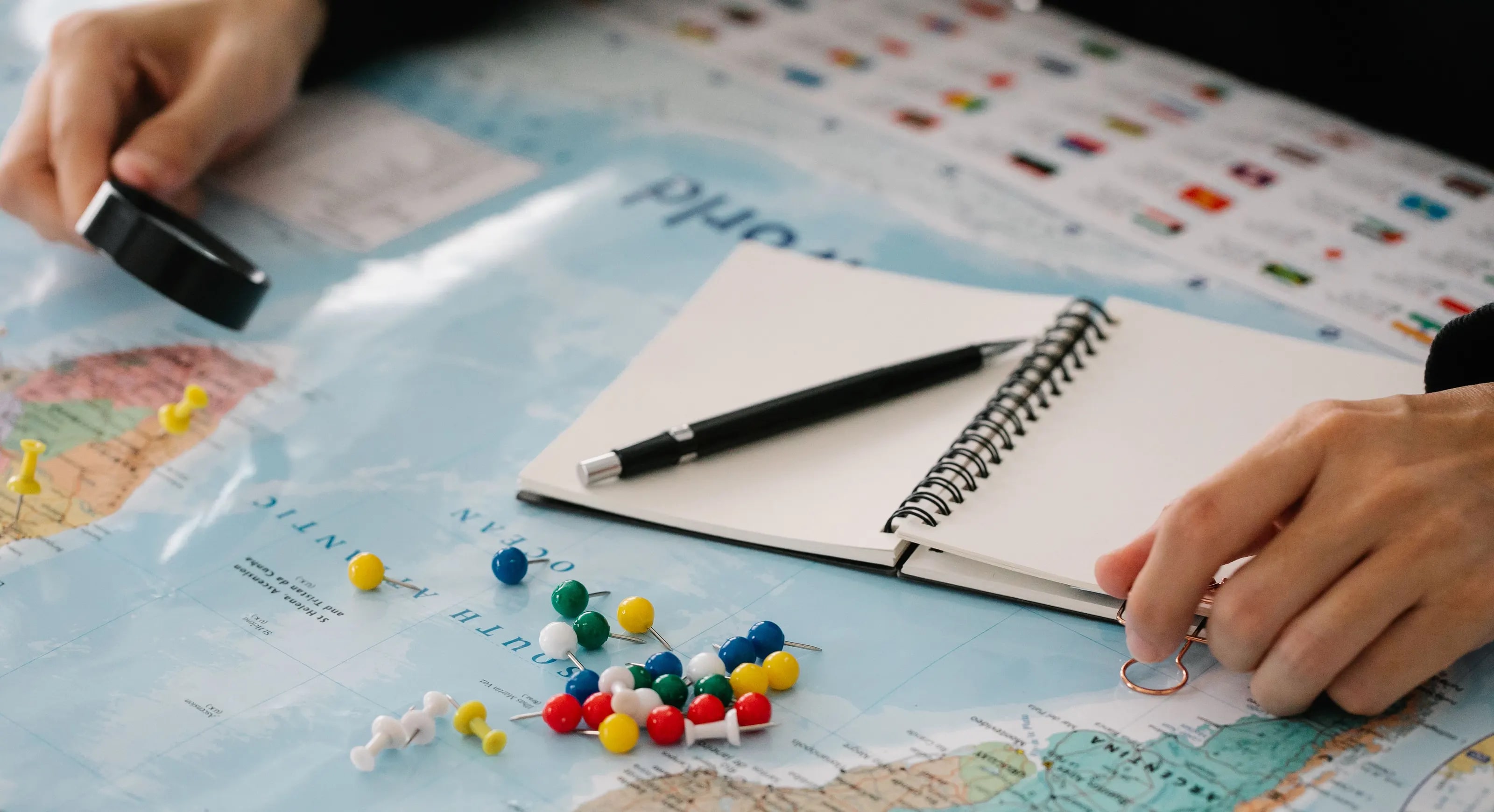 Hands at a map with coloured push pins, a notebook and a pen