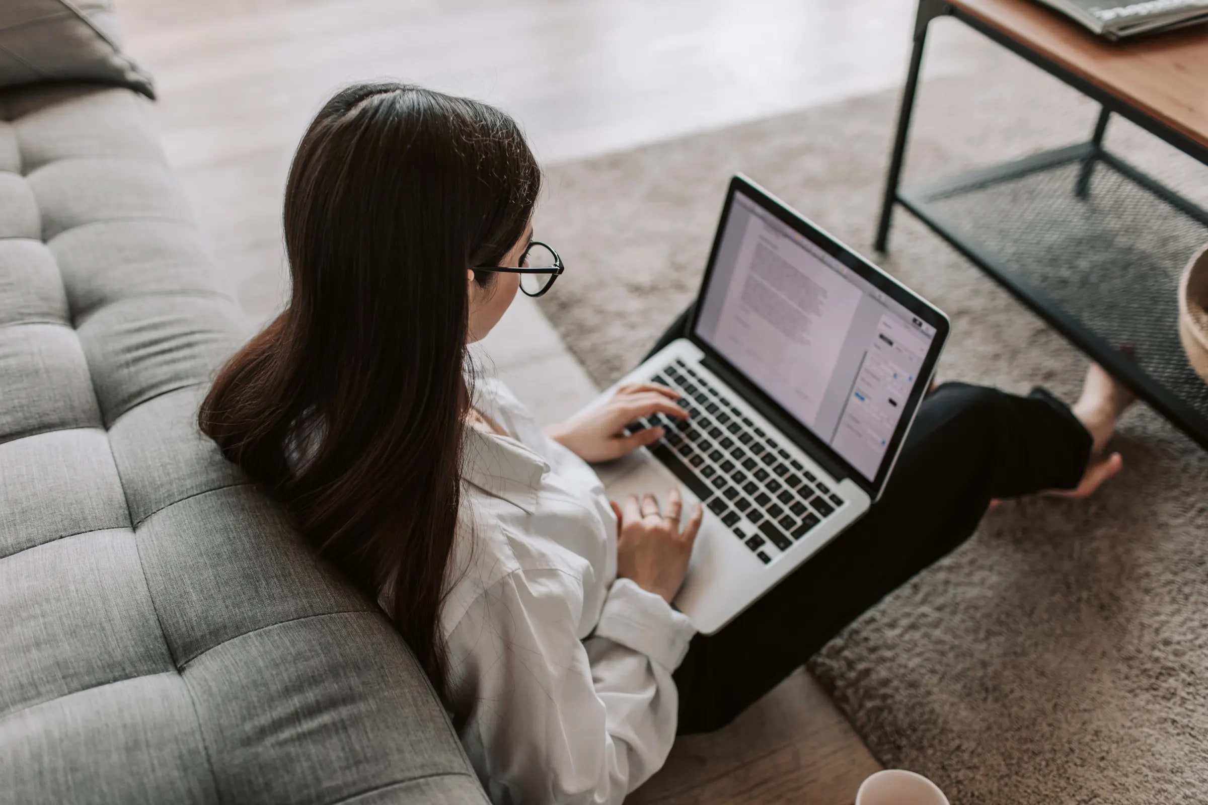A woman sits at home with a laptop on her lap, reading a document on the screen
