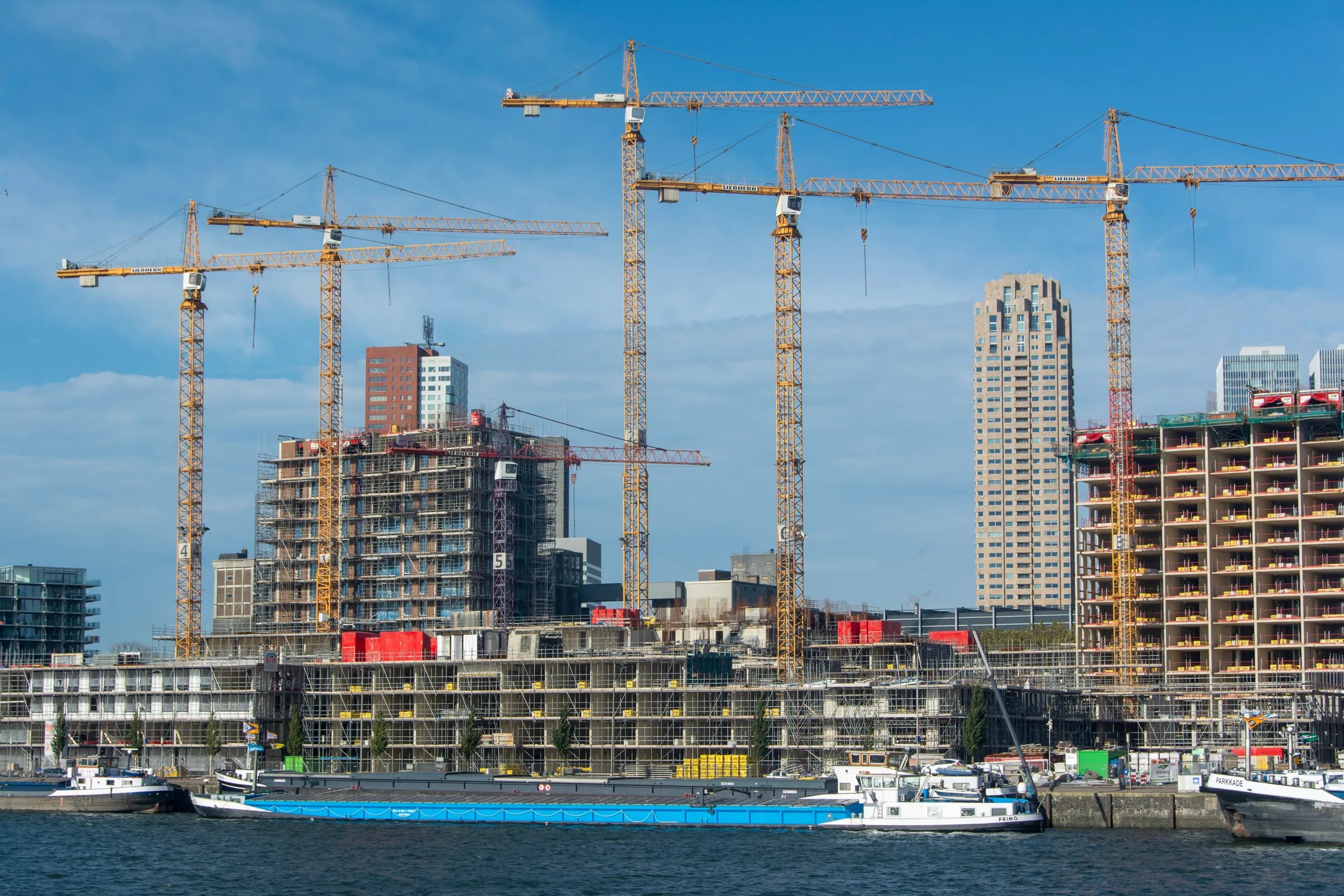Bouwkranen boven nieuwbouw aan het water, met binnenvaartschepen op de voorgrond